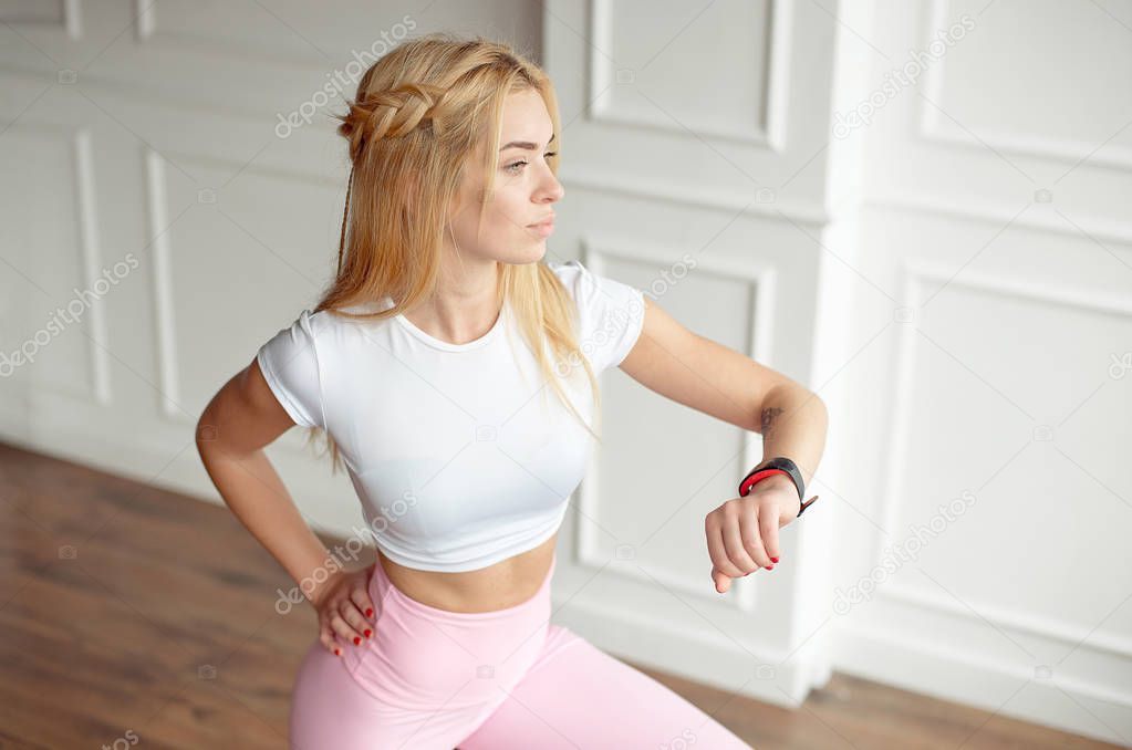 A young slim woman with an athletic body with long blond hair, wearing a white top and pink leggings in sportswear, is standing in a bright yoga room with a large panoramic window, looking at her