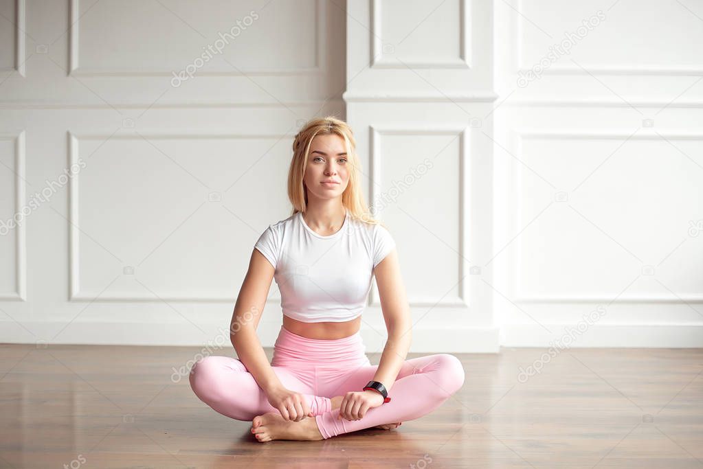 Young slim woman with an athletic body with long blond hair, dressed in white sportswear and pink leggings, sits on the floor in a bright yoga room with a large panaramic window, workout program for