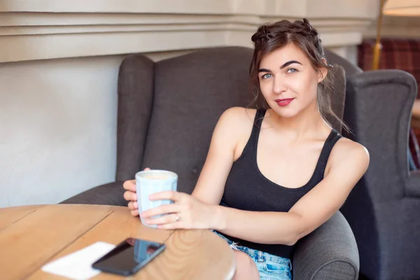 Menina atraente nova em uma camiseta preta e jeans senta-se em um café na mesa e bebe café em uma xícara vintage . — Fotografia de Stock