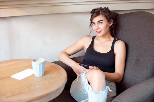 Menina atraente nova em uma camiseta preta e jeans senta-se em um café na mesa e bebe café em uma xícara vintage . — Fotografia de Stock