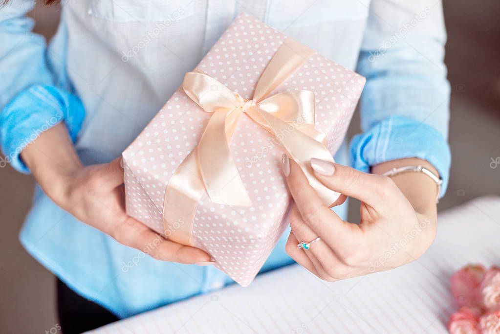 Close-up shot of female hands holding a small gift wrapped with pink ribbon. Small gift in the hands of a woman indoor.