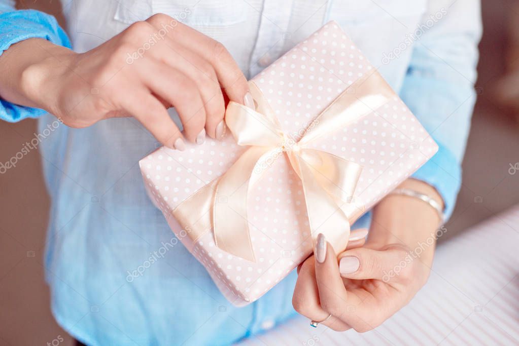Close-up shot of female hands holding a small gift wrapped with pink ribbon. Small gift in the hands of a woman indoor.