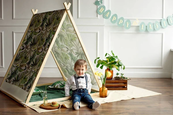 Bambino in camicia bianca, jeans e papillon è seduto in una tenda nella sua stanza decorata in stile felice Pasqua — Foto Stock
