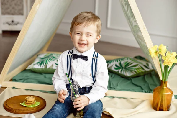 Carino bambino in camicia bianca e cravatta si siede vicino a una tenda nella sua stanza e beve limonata fatta in casa da una bottiglia e mangia biscotti fatti in casa con cioccolato — Foto Stock