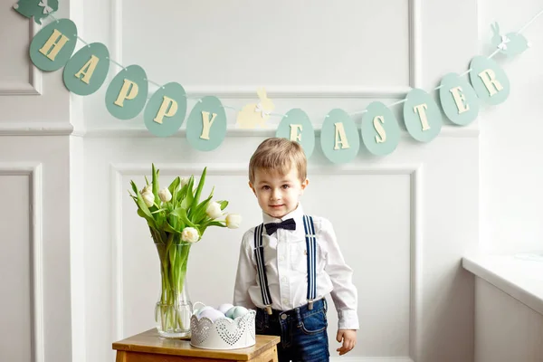 Carino ragazzo in camicia bianca e papillon è in piedi in una stanza luminosa vicino a un tavolo di legno con un vaso di vetro di tulipani e un cesto di uova di Pasqua colorate — Foto Stock