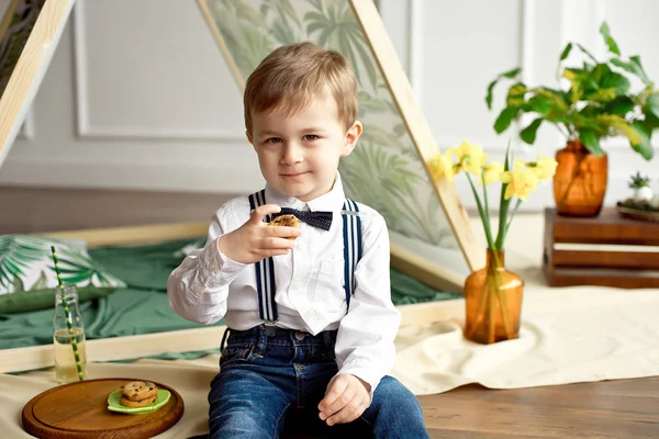 Carino bambino in camicia bianca e cravatta si siede vicino a una tenda nella sua stanza e beve limonata fatta in casa da una bottiglia e mangia biscotti fatti in casa con cioccolato — Foto Stock