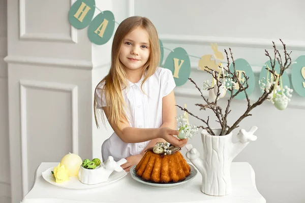 Bella bambina bionda con una camicia bianca è seduta al tavolo delle vacanze vicino a un vaso con rametti decorati con uova di Pasqua. . — Foto Stock