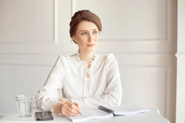 Retrato de uma bela e sorridente jovem empresária morena em uma camisa branca sentada em uma estação de trabalho moderna e brilhante trabalhando com papéis . — Fotografia de Stock