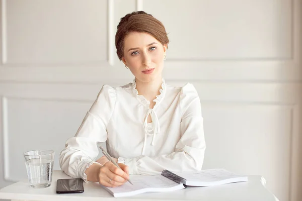 Retrato de uma bela e sorridente jovem empresária morena em uma camisa branca sentada em uma estação de trabalho moderna e brilhante trabalhando com papéis . — Fotografia de Stock