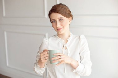 Rahat kıyafetlere çekici genç kadın, beyaz arka planda uzak ve gülümseyen bir fincan tutuyor