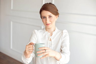 Rahat kıyafetlere çekici genç kadın, beyaz arka planda uzak ve gülümseyen bir fincan tutuyor