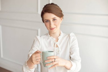 Rahat kıyafetlere çekici genç kadın, beyaz arka planda uzak ve gülümseyen bir fincan tutuyor