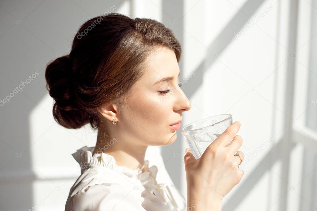 Young beautiful girl in a white shirt is standing near a window in a working office and drinking pure mineral water from a glass cup