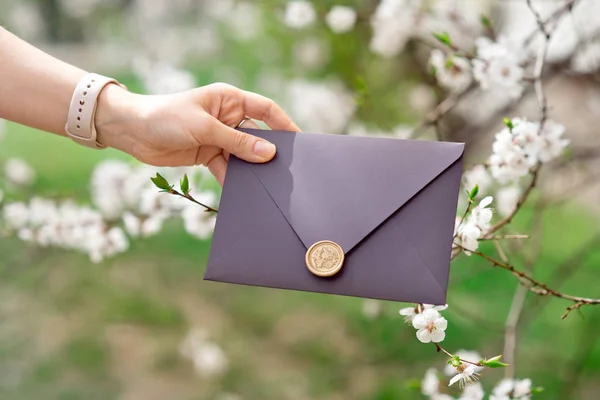 Close-up photo of female hands holding purple invitation envelope with wax seal, gift certificate,wedding invitation card on the background of blooming flowers