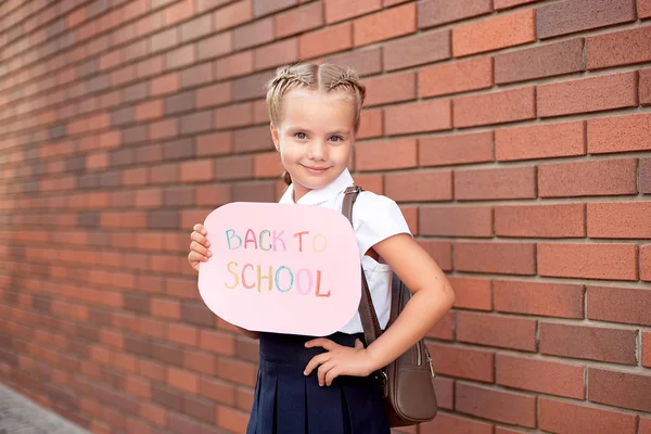 Ragazzina bionda in uniforme scolastica si trova vicino a un muro di mattoni con una lavagna con il testo torna a scuola — Foto Stock