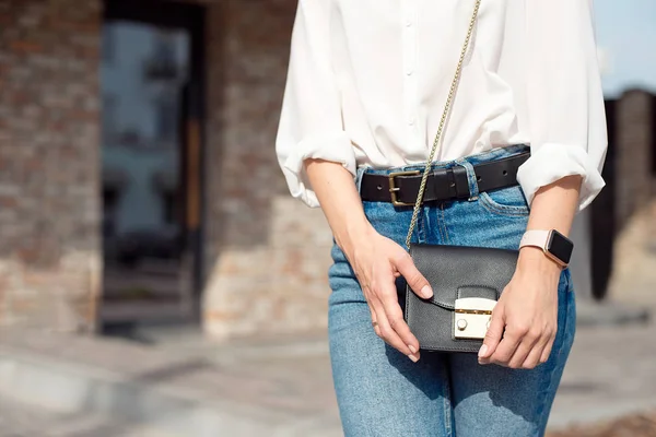 Atraktivní mladá žena v džínách a bílá košile, která stojí na ulici a drží tašku s ladymi, ve které leží její elegantní telefon a Peněženka. — Stock fotografie