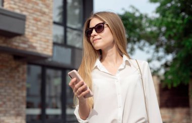 Beyaz gömlek ve patron gözlük yönetmen sevimli bir çekici çekici neşeli kız Portresi, iş merkezinin girişine yakın duran ve telefondan Sms arama.