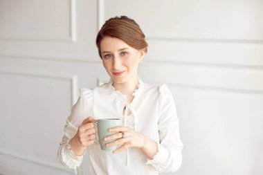Rahat kıyafetlere çekici genç kadın, beyaz arka planda uzak ve gülümseyen bir fincan tutuyor.