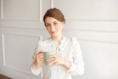 Rahat kıyafetlere çekici genç kadın, beyaz arka planda uzak ve gülümseyen bir fincan tutuyor.