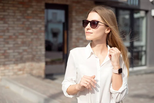 Retrato de uma mulher de negócios feliz de sucesso em uma camisa branca e óculos de sol. Relógio inteligente em uma mão fica na entrada para o centro de negócios — Fotografia de Stock