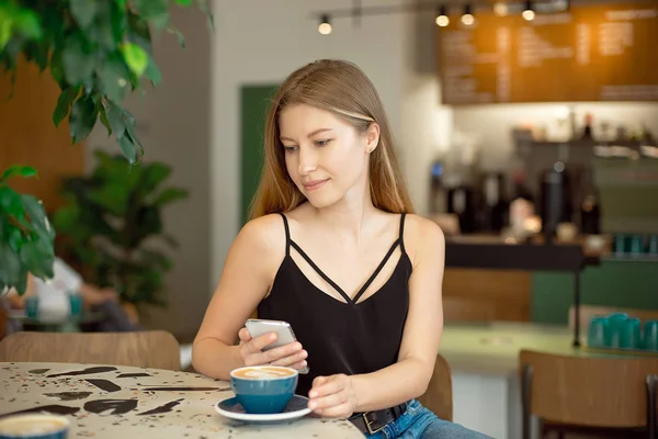 Uzun saçlı, çekici bluzlu ve kot pantolonlu genç, güzel bir sarışın kahve içer ve pencereden dışarı bakar..