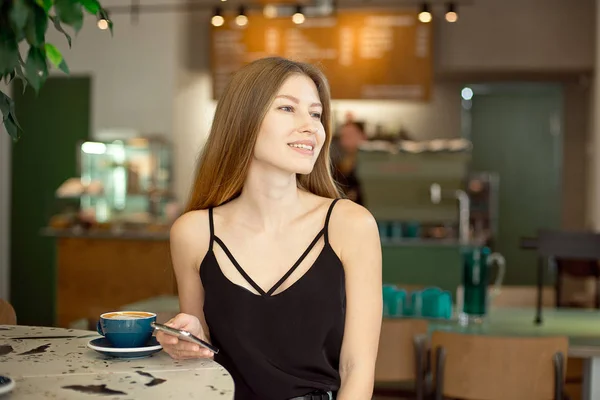 Jovem loira bonita com cabelos longos em um top charme e jeans bebe café e olha pela janela . — Fotografia de Stock