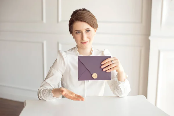 Menina em uma camisa de estilo de negócio branco com um penteado clássico no cabelo morena segurando um envelope de boas-vindas em sua mão sentado a uma mesa — Fotografia de Stock
