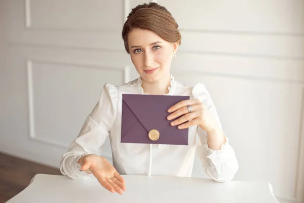 Menina em uma camisa de estilo de negócio branco com um penteado clássico no cabelo morena segurando um envelope de boas-vindas em sua mão sentado a uma mesa — Fotografia de Stock