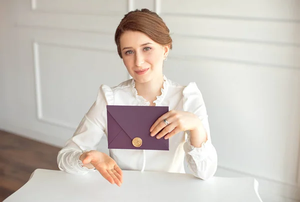 Menina em uma camisa de estilo de negócio branco com um penteado clássico no cabelo morena segurando um envelope de boas-vindas em sua mão sentado a uma mesa — Fotografia de Stock