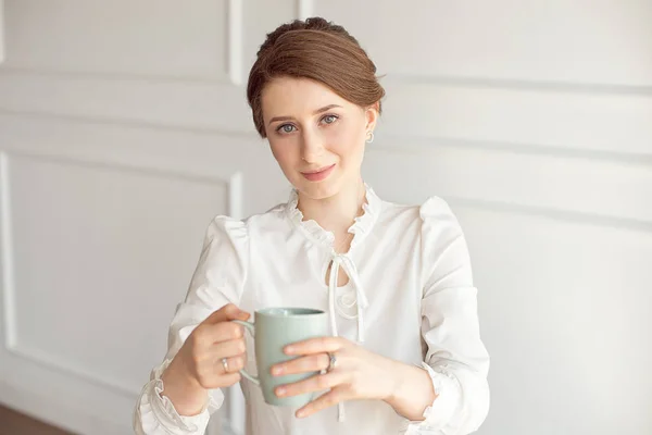 Jovem atraente em roupas casuais está segurando uma xícara, olhando para longe e sorrindo, no fundo branco . — Fotografia de Stock