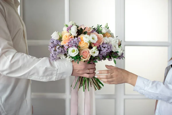 L'uomo stende le mani e dà un grande mazzo di fiori da rose e lilla isolati sullo sfondo di una vetrata. Le mani femminili di primo piano prendono un mazzo. Regalo per marzo — Foto Stock