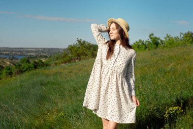 Yeşil bir çayır ve mavi gökyüzü nün arka planına karşı mutlu gülümsüyor bir hasır şapka ile uzun beyaz yazlık elbiseler giymiş büyüleyici bir pozitif kadının yarım uzunlukta portre. Güzel kadın zevk