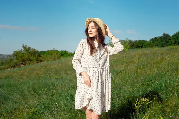 緑の草原と青い空を背景に幸せな笑顔の麦わら帽子をかぶった長い白い夏のドレスに身を包んだ魅力的なポジティブな女性の半分の長さの肖像画。美しい女性が楽しむ — ストック写真