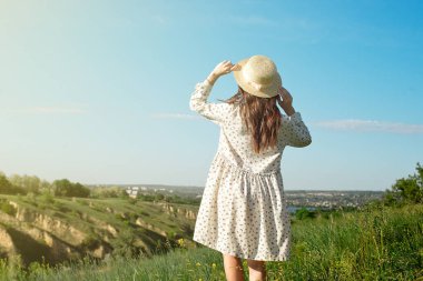 Uzun bir yazlık elbise içinde memnun esmer bir hasır şapka giyen kollarını güneş ışığına bakan kaldırdı ile uzun boylu çim duruyor.