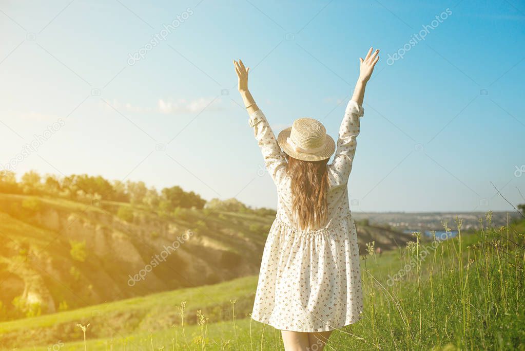 A happy woman in a straw hat with open arms stays on top of the edge of a mountain cliff under the sunset light of the sky, enjoying success, freedom and a bright future