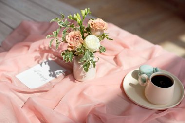 Pembe ve kahve fincanı üzerinde Fransız makaron mavi plaka çiçek gül ve yeşillik ile pembe bir masa örtüsü beyaz vazo ile ahşap bir masa üzerinde duran