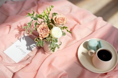 Pembe ve kahve fincanı üzerinde Fransız makaron mavi plaka çiçek gül ve yeşillik ile pembe bir masa örtüsü beyaz vazo ile ahşap bir masa üzerinde duran