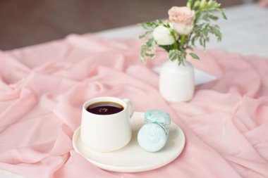 Pembe ve kahve fincanı üzerinde Fransız makaron mavi plaka çiçek gül ve yeşillik ile pembe bir masa örtüsü beyaz vazo ile ahşap bir masa üzerinde duran