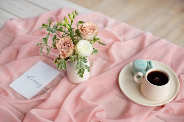 Pembe ve kahve fincanı üzerinde Fransız makaron mavi plaka çiçek gül ve yeşillik ile pembe bir masa örtüsü beyaz vazo ile ahşap bir masa üzerinde duran