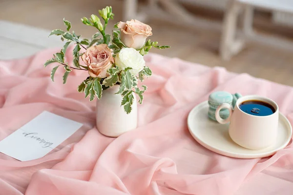 Plato azul macarrón francés en la taza de café y rosa de pie sobre una mesa de madera con un mantel rosa florero blanco con flores rosas y verdes —  Fotos de Stock