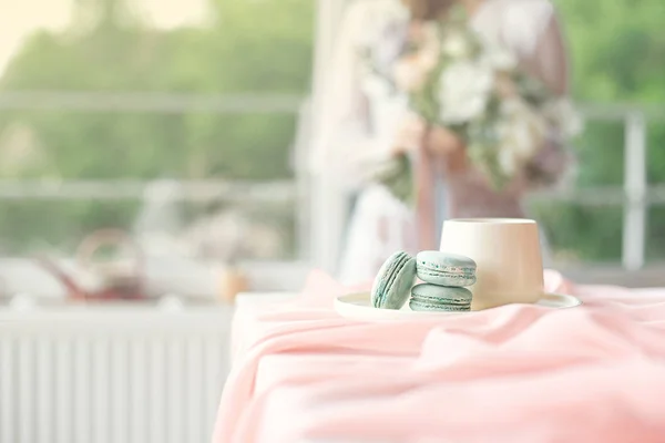 Jovem noiva caucasiana bonita desfrutando de café da manhã de macaroon francês e café em uma mesa de madeira com uma toalha de mesa rosa chiffon e um vaso de flores — Fotografia de Stock