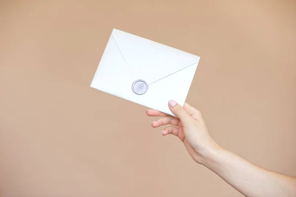 Female hands with a gentle manicure in bright colors holding an invitation to the envelope for a wedding greeting card on a beige background with a wax seal