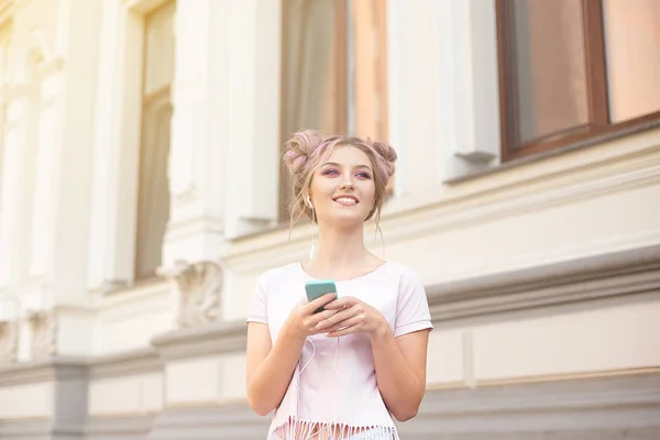 Retrato de uma jovem mulher hipster elegante andando pela rua vestida com uma roupa na moda, um penteado rosa bun viajando segurando um telefone celular — Fotografia de Stock