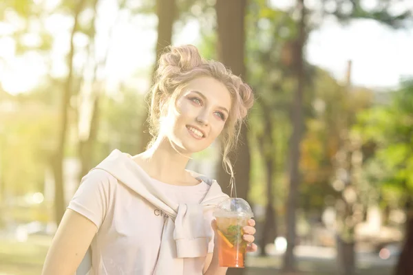 Menina bonita nova beber suco fresco de copos de plástico takeaway comida depois de uma caminhada ao ar livre. Estilo de vida saudável. Sorrindo loira magra com cabelo rosa — Fotografia de Stock