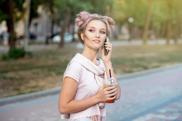 Loira jovem alegre feliz com cabelo rosa, caminha contra o pano de fundo da cidade, chama um telefone celular para seus amigos. Estudante loira está descansando na rua depois da faculdade para conhecer amigos — Fotografia de Stock