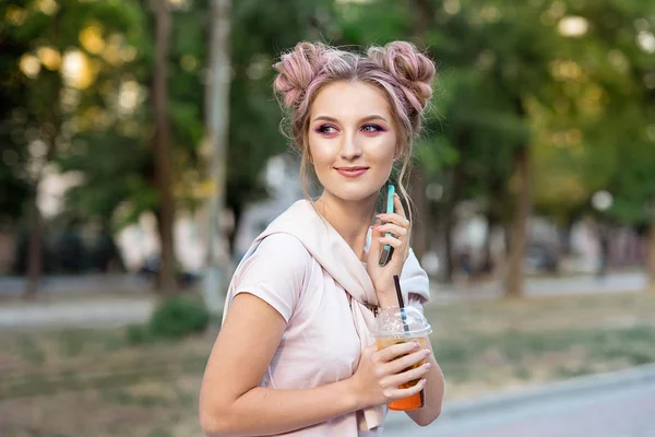 Loira jovem alegre feliz com cabelo rosa, caminha contra o pano de fundo da cidade, chama um telefone celular para seus amigos. Estudante loira está descansando na rua depois da faculdade para conhecer amigos — Fotografia de Stock