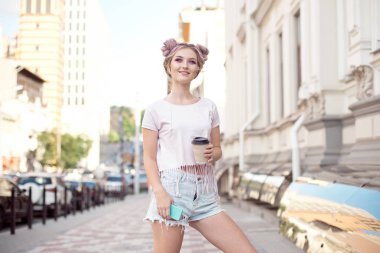 Bir saç modeli ile genç kadın bir fincan kafe tutan elleriyle telefonda kulaklıkta müzik dinlerken şehirde yürümek bir topuz ve makyaj.