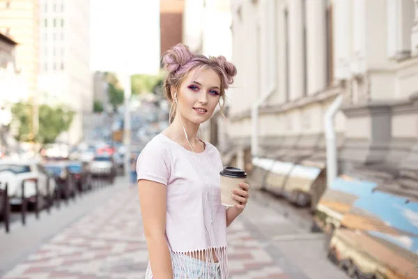 髪型のパンとメイクをした若い女性は、カフェのカップを持って彼女の手で電話を通してヘッドフォンで音楽を聴いて街を歩く. — ストック写真