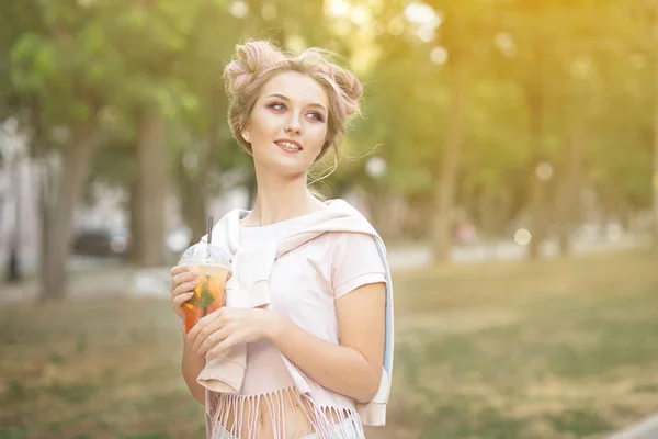 Menina bonita nova beber suco fresco de copos de plástico takeaway comida depois de uma caminhada ao ar livre. Estilo de vida saudável. Sorrindo loira magra com cabelo rosa — Fotografia de Stock