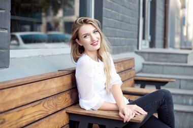 Bir açık hava restoranında bir sandalyede oturan vintage kot çekici genç kadın. Beyaz gömlekli çekici sarışın en sevdiği kafede sipariş bekliyor ve doğanın tadını çıkarıyor.